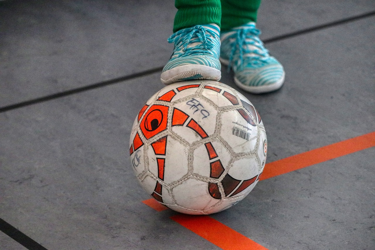 Player standing on a futsal court with one foot on a soccer ball, wearing appropriate futsal cleats.