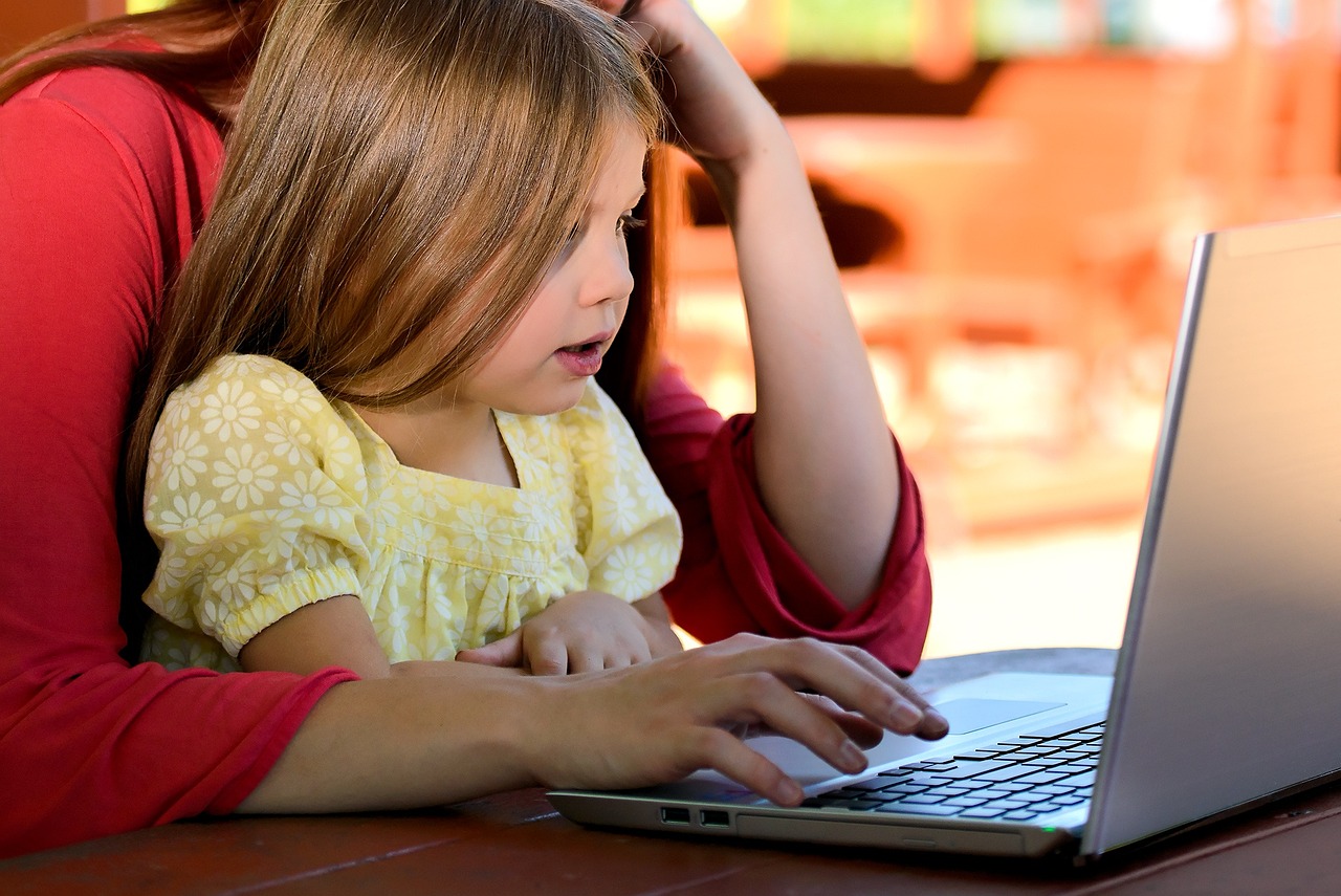 Young girl sitting on her mother's lap, both attentively looking at a laptop screen together.